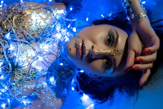 Nude girl with gold bodyart lying in the bath in blue New Year garland lights cropped close up portrait