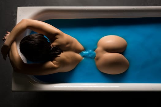 Nude brunette lying in bath in blue water high angle rearview
