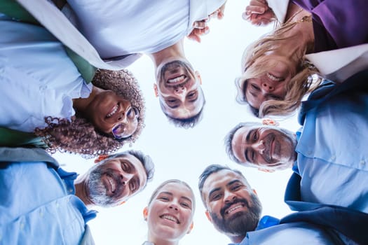 Low-angle view of a diverse group of smiling coworkers standing in a circle outdoors, symbolizing teamwork, unity, and collaboration under a clear sky.