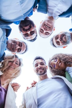 Low-angle view of a diverse group of smiling coworkers standing in a circle outdoors, symbolizing teamwork, unity, and collaboration under a clear sky.