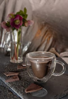 winter still life with glass transparent cup of hot chocolate on table surrounded by chocolate pieces and black chia seeds on background hellebore flowers,desert concept in color of 2025,mocha mousse