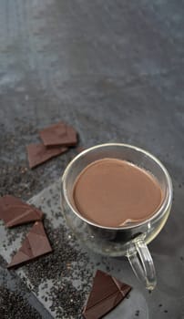 Glass cup of hot chocolate in heart shape on table surrounded by chocolate pieces and black chia seeds on grey textured background, Desert concept in color of the year 2025, mocha mousse, copy space