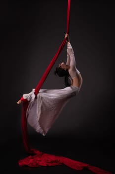 Side view of beautiful charming slim gymnast with upped hands hanging on red material on black background