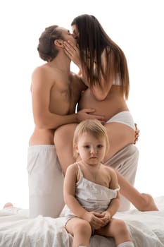 Cute baby sitting on comfortable bed in front of shirtless father kissing pregnant mother against white background