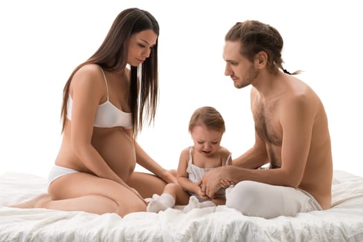 Family shot sitting on bed with their little son, father and pregnant beautiful mother isolated view