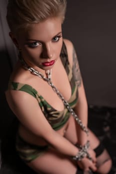 Girl in military style top sitting on the floor with a chain on her neck and wrists high angle shot in the dark room