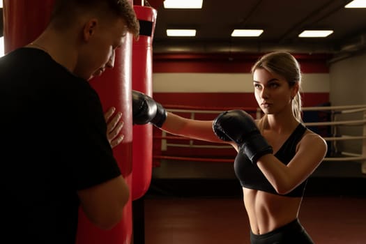 Young woman in sportswear and boxing gloves punching bag while training with male instructor in gym