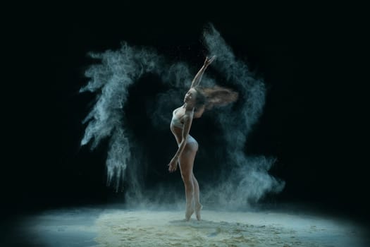 Slim girl wearing lingerie jumping gracefully in white dust cloud profile view in the dark room