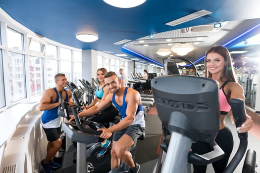 Group of cheerful young men and women smiling and looking at camera while working out on exercise machines in modern gym together