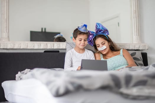 Smiling mother with bandage on her nose lies in cozy bed and watches TV together on tablet, mother has big blue bow on her head and son has blue hat, carnival in bed to lift mood, relax,