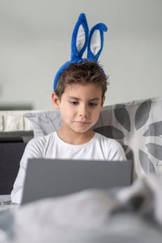 Boy with blue bunny ears sitting on sofa, concentrating on working on laptop, Casual atmosphere, playful and cheerful mood before Easter,