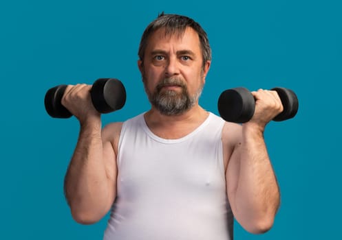 An elderly man lifts dumbbells while focusing on strength training. He wears a sleeveless top and exercises in a well-lit gym environment, showcasing dedication to fitness.