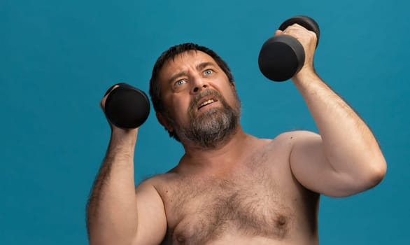 An elderly man, shirtless and focused, lifts dumbbells above his head in a bright, uncluttered space. His expression shows determination as he engages in strength training.