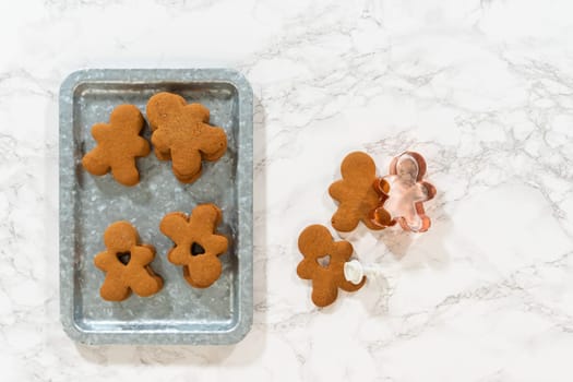 Flat lay. Gingerbread cookies, including a gingerbread man with a heart-shaped cutout, rest on a rustic metal tray against a marble countertop.