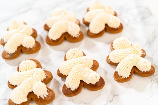 Gingerbread cookies await their second halves on a marble surface, each meticulously piped with buttercream to craft delightful sandwich treats. The precision of the piping adds a festive touch.