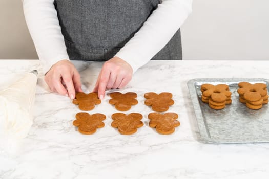 Gingerbread cookies await their second halves on a marble surface, each meticulously piped with buttercream to craft delightful sandwich treats. The precision of the piping adds a festive touch.