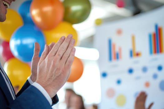 A close-up of hands clapping in a professional setting, surrounded by colorful balloons and informative charts, depicts a festive and motivational atmosphere.