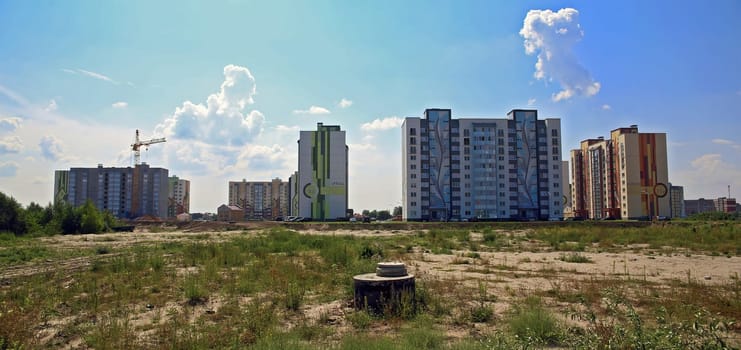 Tall residential structures are visible in a developing urban area with a construction crane in the background. The landscape features sparse greenery, indicating an evolving neighborhood during daytime.