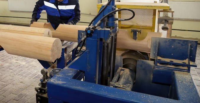 A worker efficiently uses a machine to cut and shape logs in a well-lit workshop, surrounded by wood and tools.