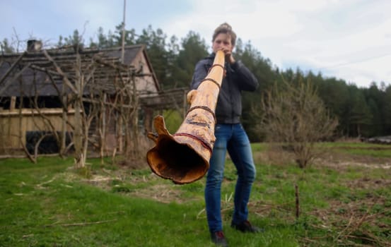 A person is showcasing a traditional wind instrument crafted from bark in a natural setting filled with greenery and rustic structures.
