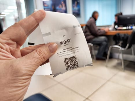 Kirov, Russia - October 08, 2024: A person holds a ticket with queue number F047 while waiting in a busy bank on a typical weekday.