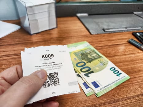 Kirov, Russia - October 08, 2024: Hand holds a currency exchange voucher beside a stack of euros on a wooden surface at a financial establishment.
