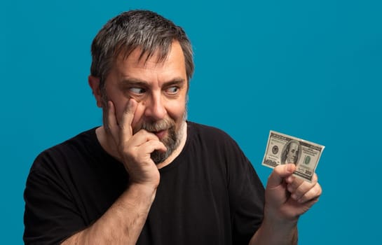A middle-aged man with a beard holds a hundred dollar bill while looking contemplative. His hand rests on his face, suggesting he is pondering an important decision or idea.