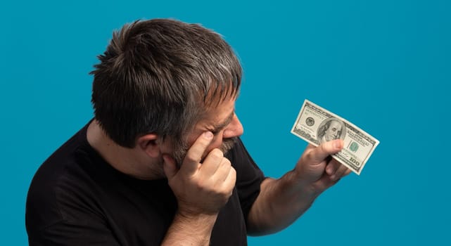 A man is closely inspecting a one hundred dollar bill, demonstrating curiosity and focus. His expression hints at interest while he holds the bill against a vivid blue backdrop.