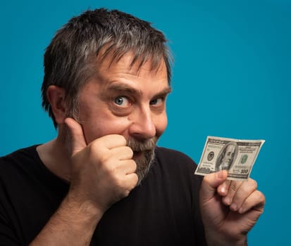 A man with a beard and short hair expresses joy by giving a thumbs up while holding a one hundred dollar bill. The bright blue background adds to the cheerful atmosphere, highlighting his excitement.