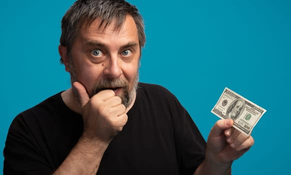 A middle-aged man with a gray beard expresses excitement, holding a hundred dollar bill while giving a thumbs up. The vibrant blue background enhances his enthusiastic demeanor.