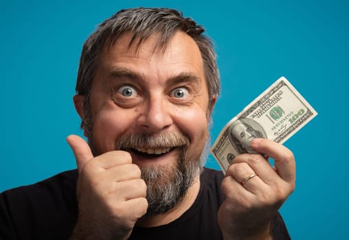 A smiling man with a beard enthusiastically displays a dollar bill. He gives a thumbs up, expressing joy and satisfaction against a bright blue backdrop.