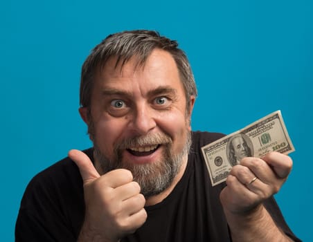 A man with a joyful expression holds a hundred-dollar bill in one hand while giving a thumbs-up with the other. The bright blue background enhances his enthusiastic demeanor.