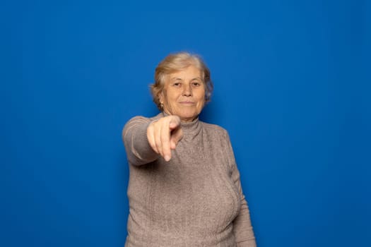 Caucasian woman in her 70s wearing a dark beige sweater pointing at the camera while smiling kindly, isolated over blue studio background