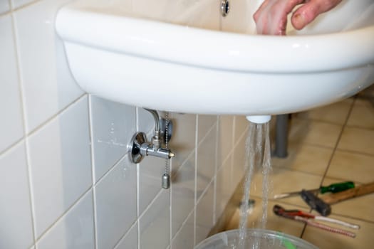 Water flows from a white sink into a bucket in a bright bathroom with tiled walls, concept of repairing pipes and replacing plumbing,