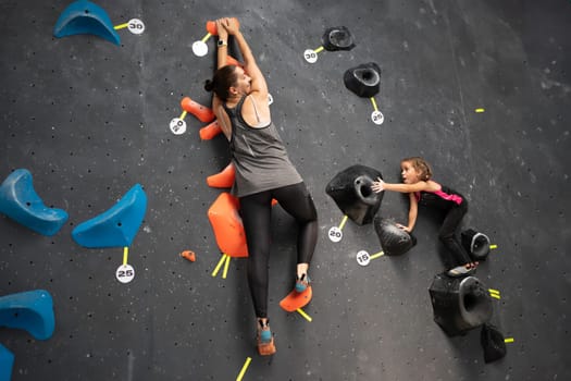 Determined mother and daughter climbing artificial rock wall at bouldering gym. From below of sporty female athletes are training on equipment.