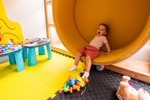 Happy child sitting inside a round yellow play structure in a colorful playroom filled with activity tables and toys for learning and fun