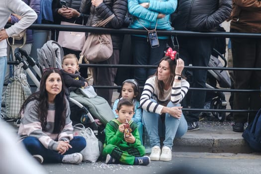 Family-friendly event spectators: Two mothers and three kids, one in a stroller, behind barriers at a public gathering. Crowded background indicates festive occasion.