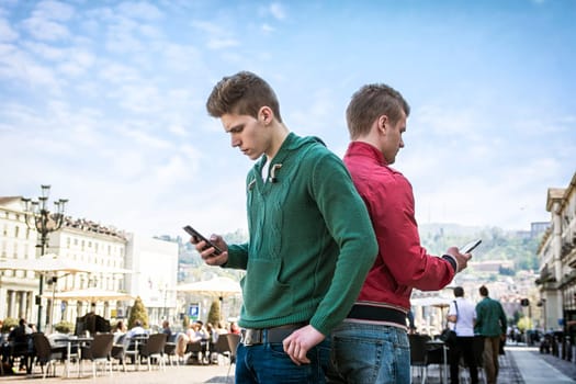 A couple of men standing next to each other, back to back, engrossed in their cell phones and ignoring each other's company