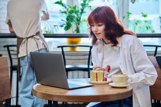 Female college student with laptop in cafeteria at table with cup of coffee and piece of cake. Internet online technology for leisure communication blogging learning chat, youth lifestyle concept