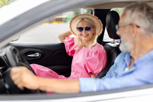 Smiling excited senior couple husband and wife going summer vacation by car together. Happy retired man and woman wearing wicker hat and sunglasses sitting in auto, enjoying road trip, chatting
