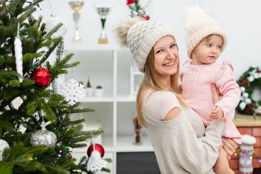 A smiling mother and her little daughter spend time together during the holiday season. A white bookcase with shelves can be seen in the background. A decorated Christmas tree stands next to the woman and her child.