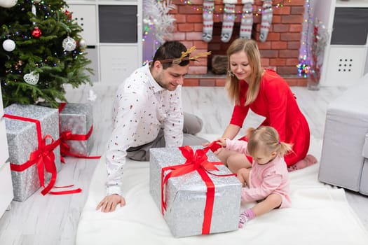 The woman is dressed in a red dress. A woman, a man and a little girl are sitting on the floor. The family is sitting around a large Christmas present. A section of a Christmas tree can be seen next to the family.