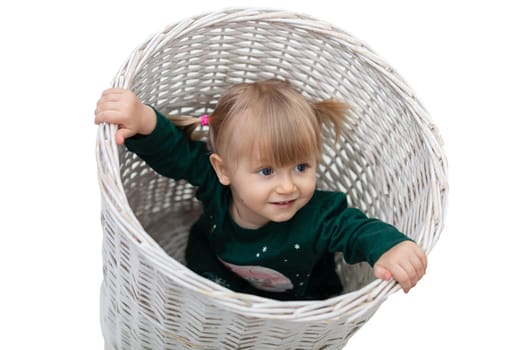 The young child is wearing a dark green tracksuit set. The girl is combed into two ponytails. The child is playing, sitting in a large white wicker basket. White background.