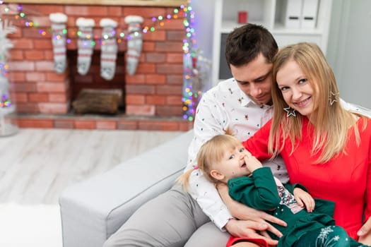 A man, a woman and a little girl sit together on a gray couch. The woman holds her lying daughter in her lap. The blurry background shows a brick fireplace decorated with Christmas lights.