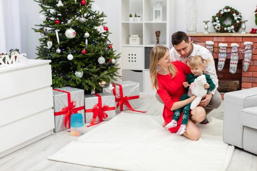 Under the decorated Christmas tree lie large gifts tied with red ribbons. Next to the Christmas tree is a man, a woman and a little girl. The mother is hugging her little daughter. The family members smile at each other.