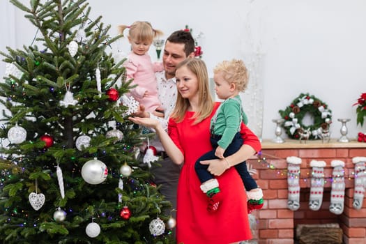 Mom and dad are holding their children in their arms. The man is holding a little girl and the woman is holding a little boy. Parents stand with their children by the Christmas tree. Parents show the Christmas tree to their children.