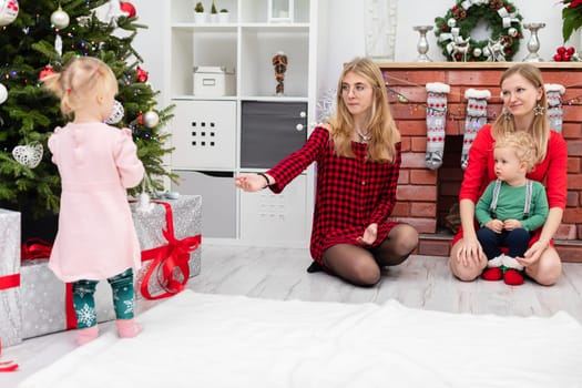 Two young women are kneeling by the fireplace. Both are blonde. One of the women wears glasses. There are also children in the room: a little girl and a little boy. The little girl is standing by the Christmas tree, under which there are presents.