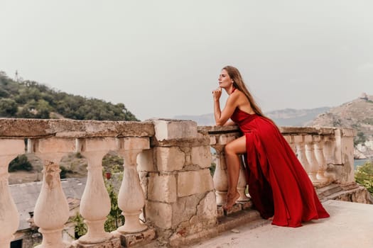 Side view a Young beautiful sensual woman in a red long dress posing on a volcanic rock high above the sea during sunset. Girl on the nature on overcast sky background. Fashion photo