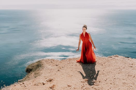 Side view a Young beautiful sensual woman in a red long dress posing on a rock high above the sea during sunrise. Girl on the nature on blue sky background. Fashion photo.