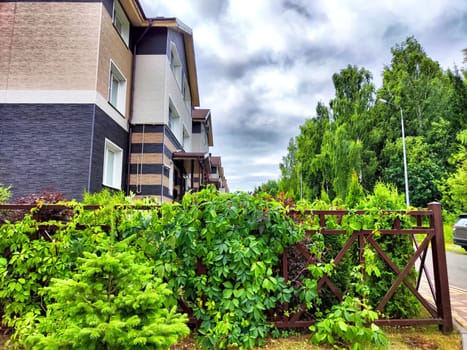Lush greenery envelops a wooden fence near modern homes under a cloudy sky.
